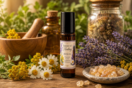 An amber glass roll-on bottle labeled 'Essence of Healing' botanical Bruise oil stands on a rustic wooden surface, surrounded by lavender, chamomile flowers, crystals, a wooden mortar and pestle, and glass jars in a warm, natural setting.