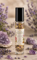 A clear glass roller bottle filled with calming oil and dried lavender flowers, topped with a black cap. The bottle is centered on a beige surface with decorative purple crystals and dried lavender sprigs around it.