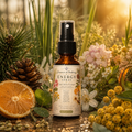 An amber glass spray bottle labeled 'Essence of Healing Energy Spray' stands centered among natural props including pine needles, a pinecone, dried orange slices, green leaves, and yellow flowers in a sunlit outdoor-style setting.