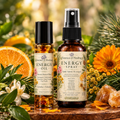 Two botanical wellness products from Essence of Healing, Energy Oil and Energy Spray, displayed on a rustic wooden surface surrounded by fresh orange slices, white flowers, yellow flowers, pine needles, and crystals against a soft, blurred natural background.