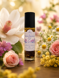 A bottle of Grounding Oil by Essence of Healing stands on a wooden surface surrounded by pink roses, white and yellow flowers, with a warm, softly blurred background.