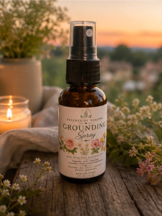 A bottle of Essence of Healing Grounding Spray stands on a rustic wooden surface outdoors at sunset, surrounded by flowers, a lit candle, and soft natural background blur.