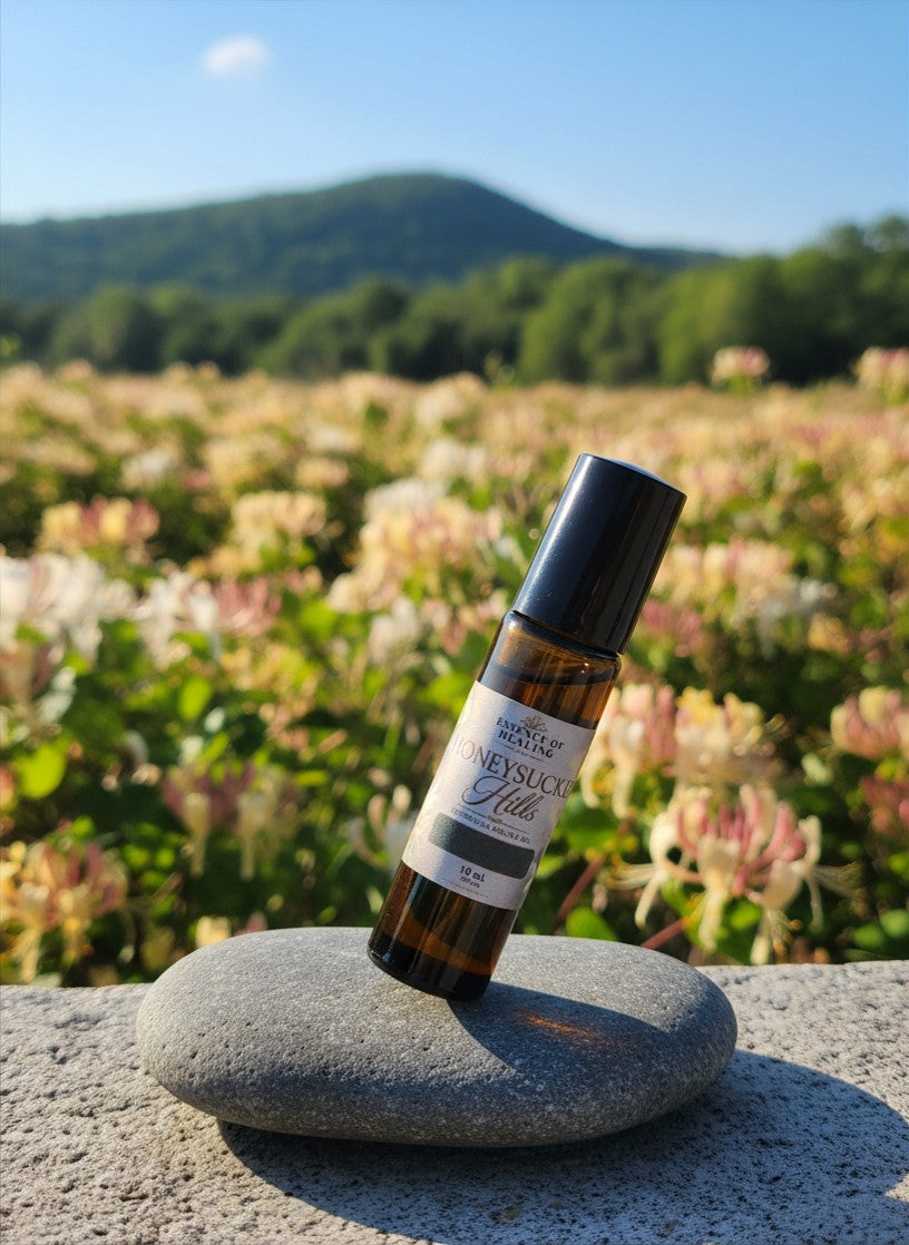 A small amber glass roll-on bottle with a black cap and a white label Honeysuckle Hills scented body oil stands on a smooth gray stone in front of a blurred field of pale pink flowers and green hills under a blue sky.