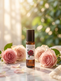 A small amber glass roller bottle labeled Rose Essence'ESSENCE OF HEALING' stands centered on a marble surface, surrounded by pink roses and soft green foliage in a bright, sunlit setting.