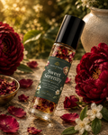 A clear glass roller bottle of Sweet Serenity scented body oil with a black cap, surrounded by red roses, petals, and white blossoms on a stone surface with a warm blurred garden background.