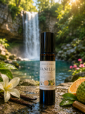 Vanilla Woods essential oil bottle with vanilla beans, oranges, and flowers in front of a waterfall.